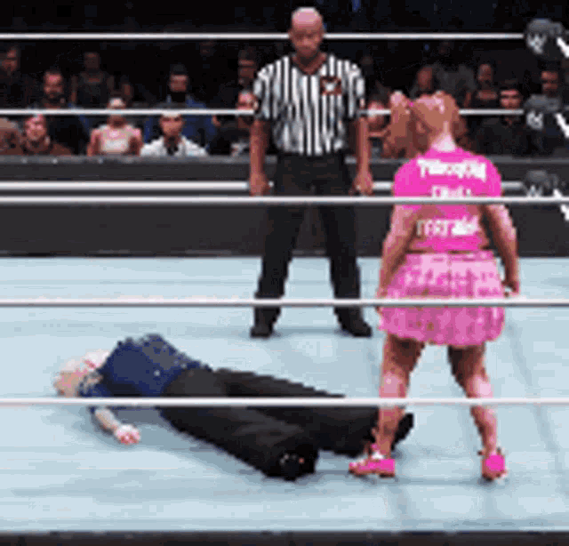 a woman in a pink skirt stands next to a man laying on the ground in a wrestling ring