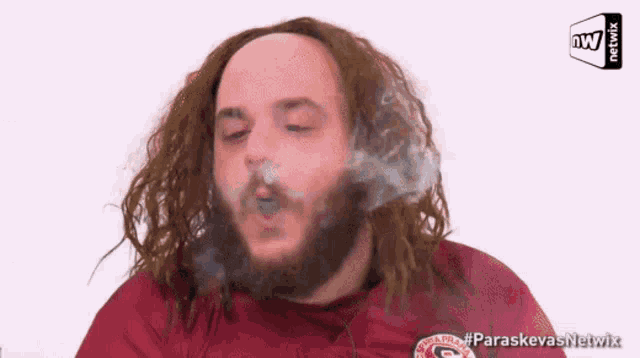 a man with long hair and a beard is blowing smoke out of his mouth while wearing a paraskevas netwix shirt