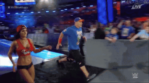 a man in a blue shirt and a woman in a red bikini are running in a wrestling ring .
