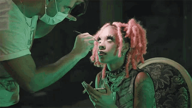 a woman with pink hair is getting her makeup done by a man wearing a face mask