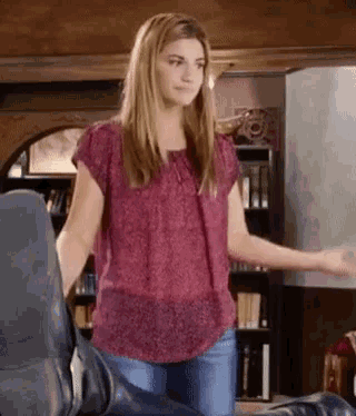 a woman in a pink shirt and blue jeans is standing next to a chair in a room .