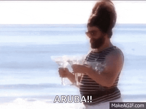 a man with a beard is standing on a beach holding a martini and says aruba !