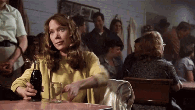 a woman is sitting at a table holding a glass and a bottle of coke