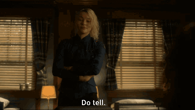 a woman in a blue shirt stands in front of a window with blinds and says do tell