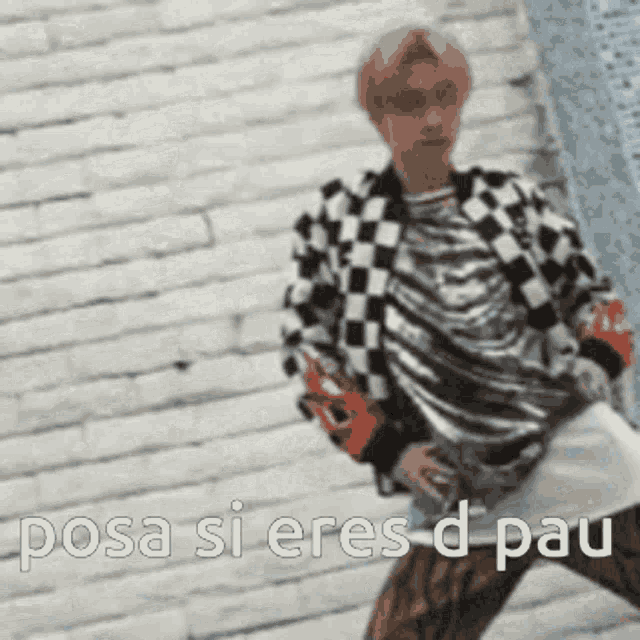 a man in a checkered jacket is standing in front of a brick wall and says `` posa si eres d pau '' .
