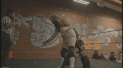 a referee watches two women wrestle in a ring with a mural on the wall behind them with the number 25 on it