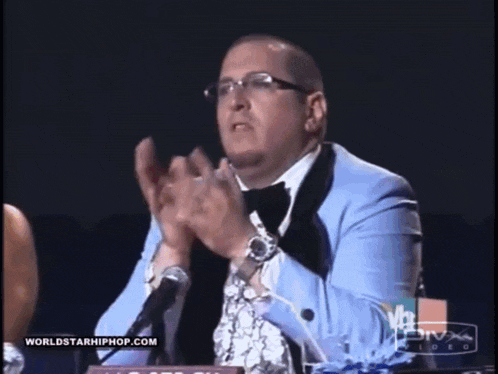 a man in a tuxedo applauds in front of a worldstarhiphop.com sign