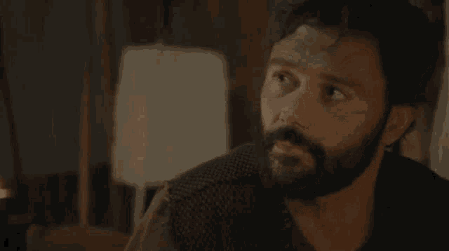 a man with a beard is sitting at a table with a lamp in the background .