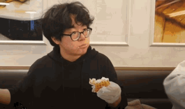 a man wearing glasses and a black hoodie is holding a piece of food in his hand .