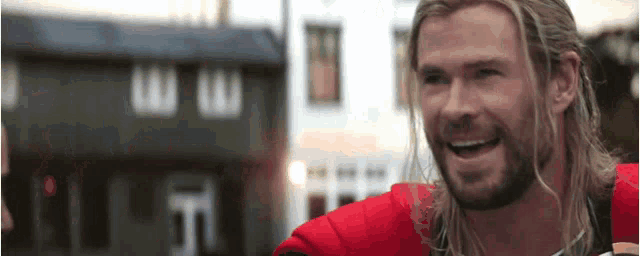 a man with long hair and a beard is smiling in front of a building in a city .