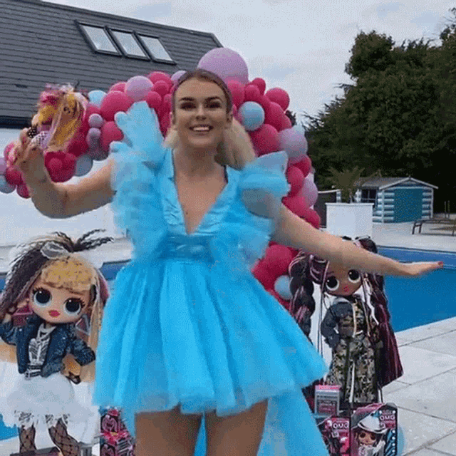 a woman in a blue dress is standing next to a pool holding balloons and dolls