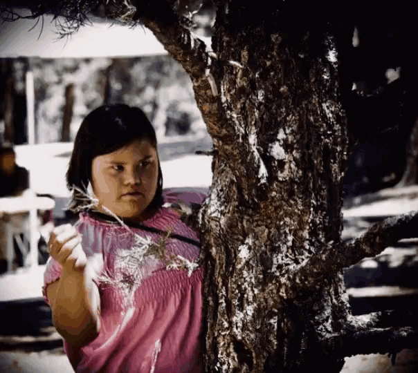 a young girl in a pink shirt is standing next to a tree