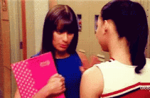 two cheerleaders are talking to each other in a locker room while one is holding a pink folder .