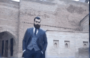 a man with a beard wearing a suit and tie is standing in front of a brick building .
