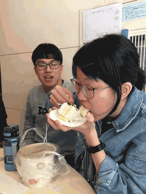 a woman eating a piece of cake with a man behind her wearing a sweatshirt with the letter k on it
