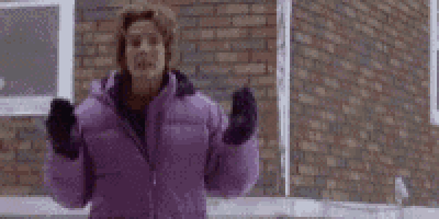 a woman in a purple jacket and mittens is standing in front of a brick building .