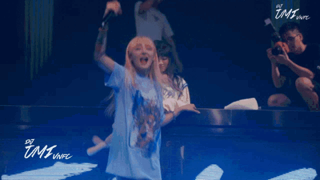 a woman in a blue t-shirt is dancing on a stage with the words dj tmi vnfc written on it