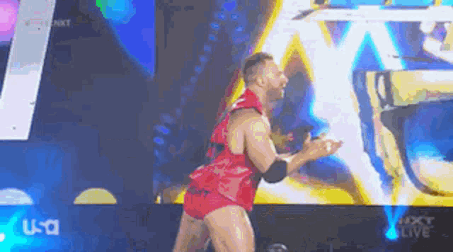 a wrestler in a red shirt and shorts is standing on a stage in front of a large screen .