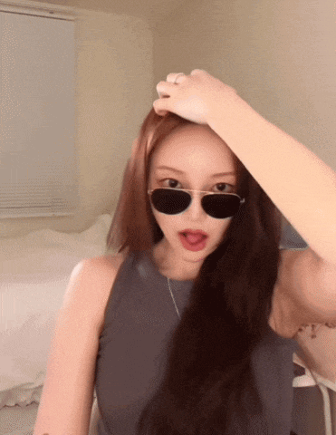 a woman wearing sunglasses and a gray tank top holds her hand to her head