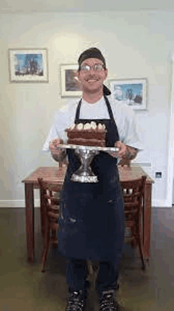 a man in an apron is standing in front of a table holding a cake on a plate .
