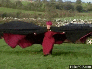 a man in a red and black cape is standing in a field holding a black cloth .