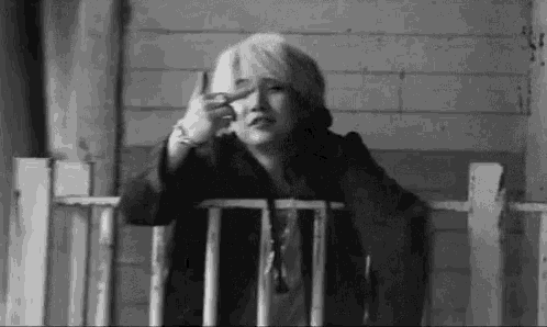 a black and white photo of a woman sitting on a porch making a peace sign with her hand .