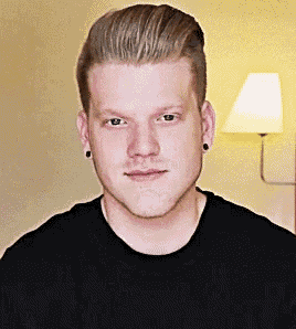 a young man wearing a black shirt and earrings looks at the camera in front of a lamp .