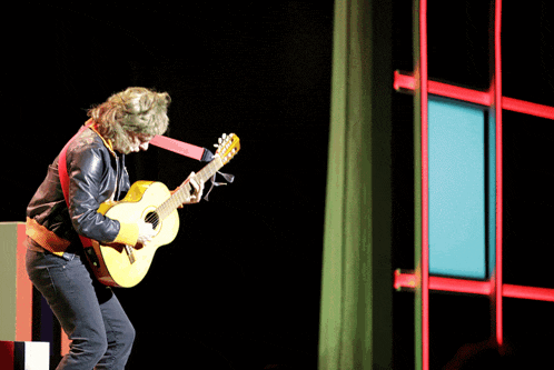 a man is playing a guitar on a stage with a green curtain behind him