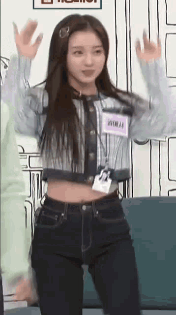 a woman in a crop top and jeans is standing on a couch with her arms in the air .