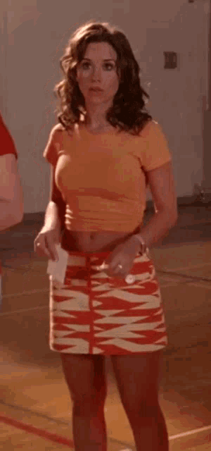 a woman wearing an orange crop top and an orange and white skirt is standing in a gym