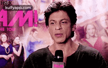 shah rukh khan is talking into a microphone in front of a poster of a movie .
