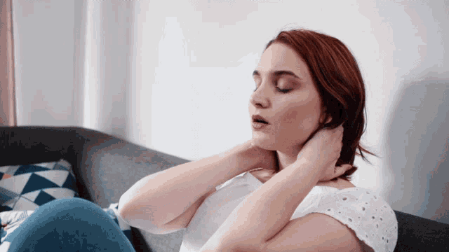 a woman sits on a couch with her eyes closed and her hands on her neck
