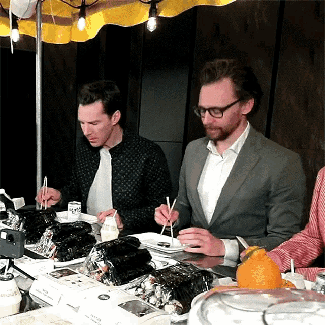 two men sitting at a table eating food with chopsticks