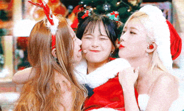 three girls are wearing santa hats and costumes and kissing each other on the cheeks .