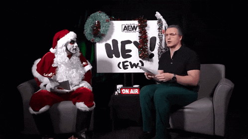 a man in a santa suit is sitting next to a man in a black shirt with the words hey you behind the camera
