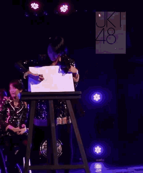 a woman is sitting at a table in front of a sign that says jk48 on it .