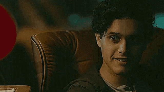a young man with curly hair is sitting in a chair and smiling .
