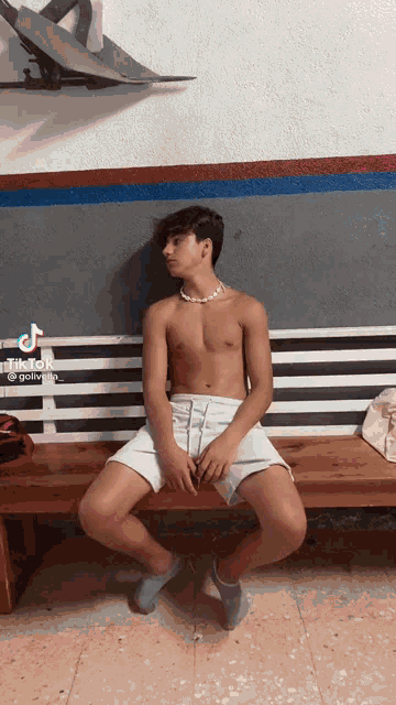 a shirtless young man is sitting on a wooden bench with his legs crossed .