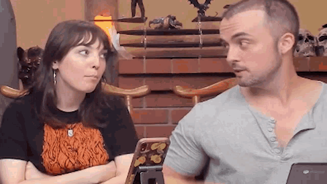 a man and a woman are sitting next to each other in front of a fireplace and looking at each other .