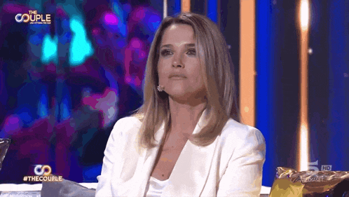 a woman in a white jacket is sitting in front of a screen that says " the couple "