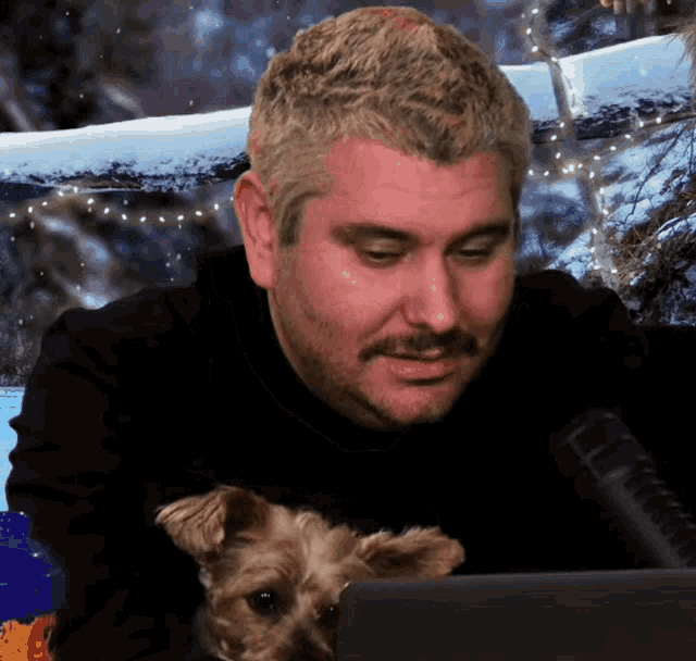 a man with a beard and mustache is holding a small dog while looking at a laptop
