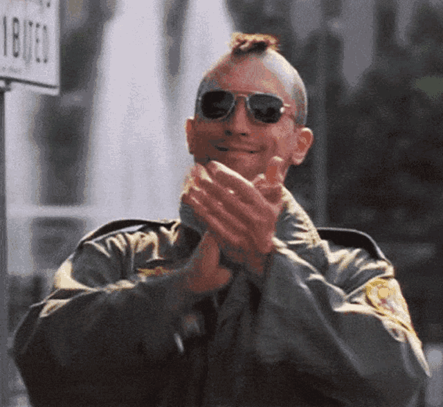 a man wearing sunglasses and a mohawk applauds in front of a sign that says ' driven '