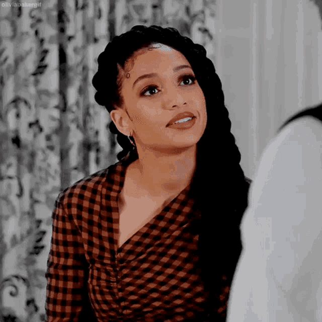 a woman with braids is wearing a plaid shirt and earrings and talking to a man .