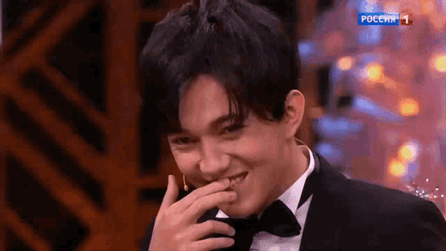 a man in a tuxedo and bow tie is smiling and covering his mouth with his hand while on a tv show in russia