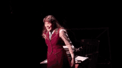 a woman in a red dress is standing on a stage in front of a desk with papers on it .