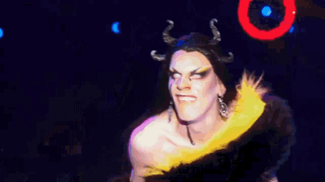 a drag queen with horns and feathers says " you " at the bottom