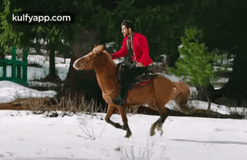 a man in a red jacket is riding a brown horse through the snow .