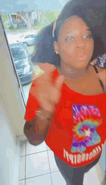 a woman is standing in front of a window wearing a red tie dye shirt .