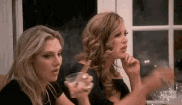 two women are sitting at a table holding drinks and talking .