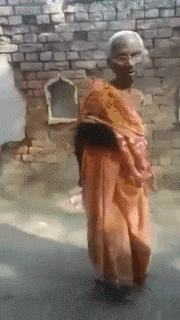 an elderly woman in an orange sari is standing in front of a brick wall